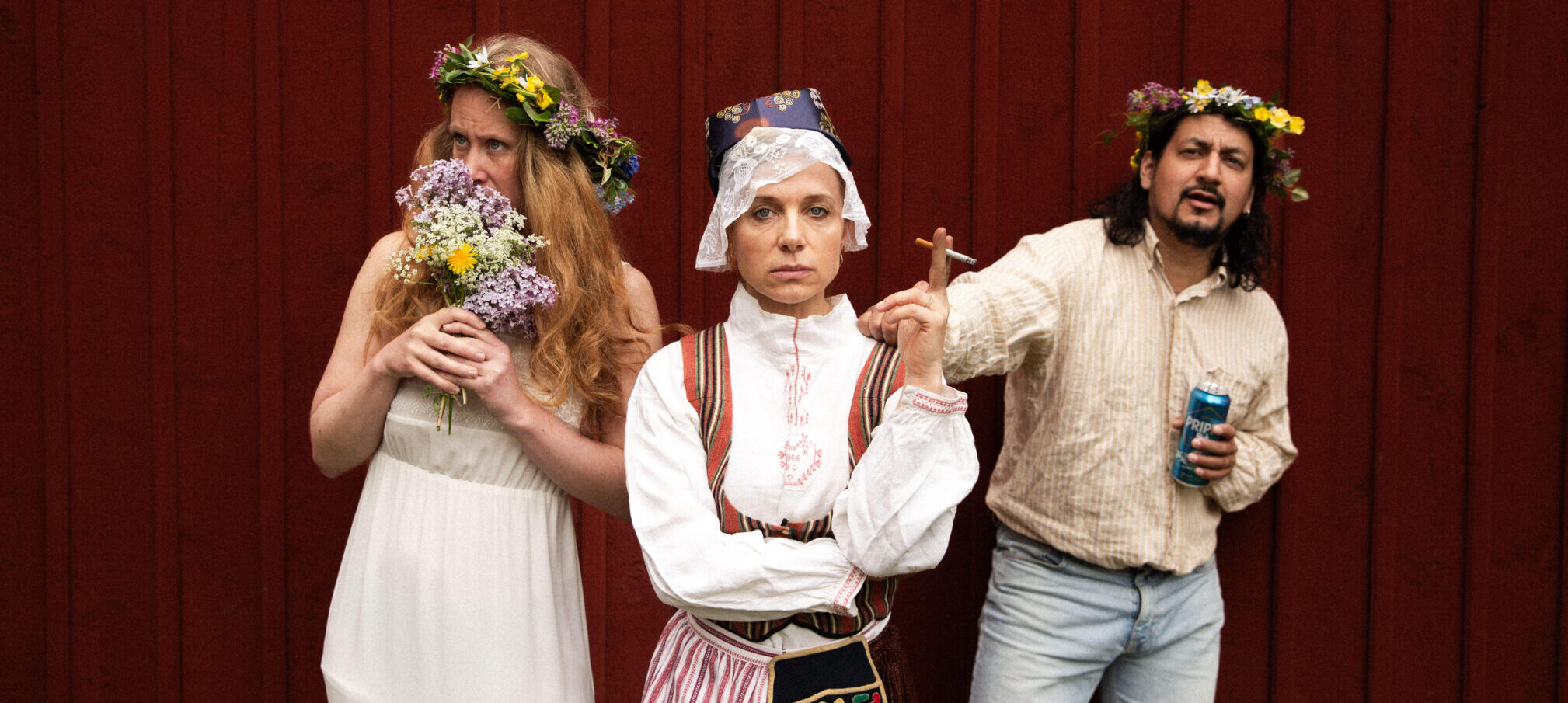 Three people in midsummer outfits in front of a red wooden wall. One smells flowers, one wears traditional folk costume, one looks slightly drunk.