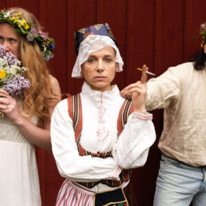 Three people in midsummer outfits in front of a red wooden wall. One smells flowers, one wears traditional folk costume, one looks slightly drunk.