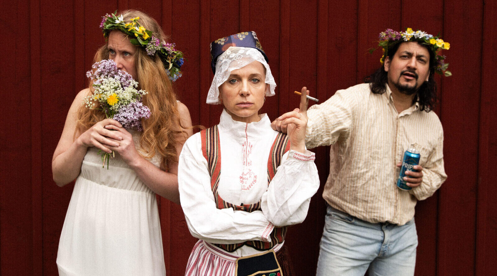 Three people in midsummer outfits in front of a red wooden wall. One smells flowers, one wears traditional folk costume, one looks slightly drunk.