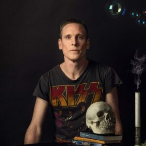 En medelålders man i en kiss-tshirt framför ett bord med en döskalle och ett stearinljus
