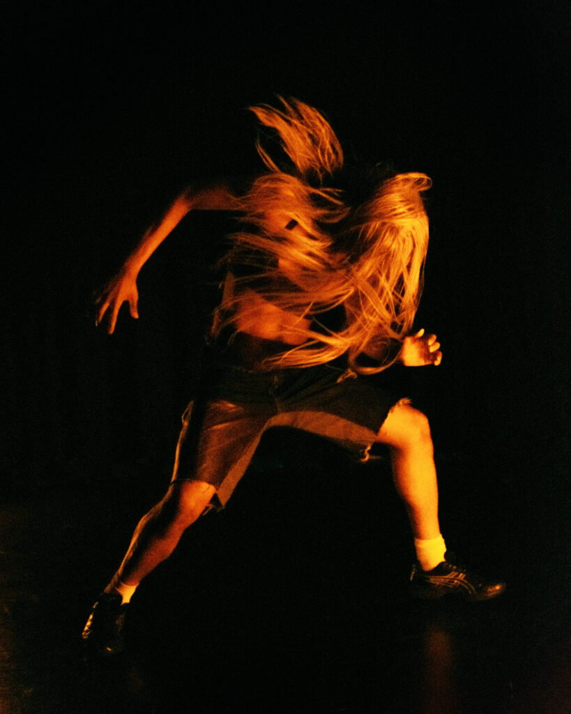 En man med långt hår dansar i sidljus på en scen.